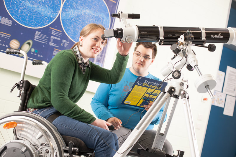 Schülerin im Rollstuhl schaut durch ein Teleskop. Daneben sitzt ein Schüler mit Sternenkarte in der Hand.