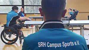 Das SRH-SHS-Rollstuhl-Tischtennis-Team beim Tischtennisspiel; im Vordergrund junger Mann mit SRH-Campus-Sport-T-Shirt